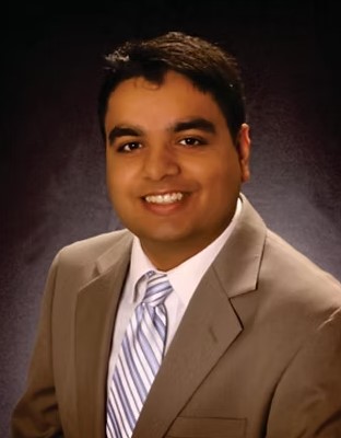 The image shows a person wearing a beige suit and a striped tie, smiling against a dark background.