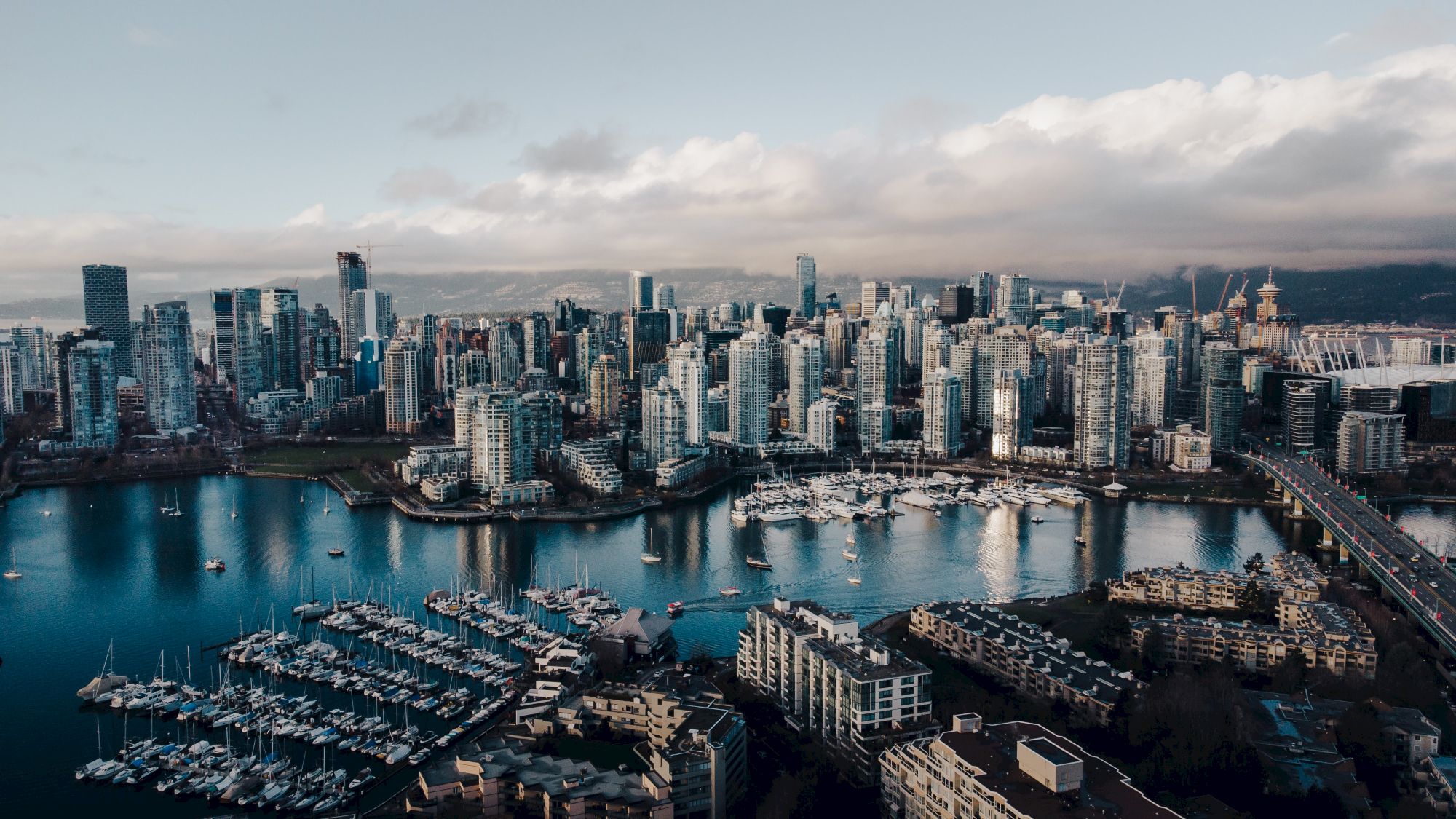 Aerial view of a modern city skyline with tall buildings, a marina with boats, and a bridge over water under a partly cloudy sky.