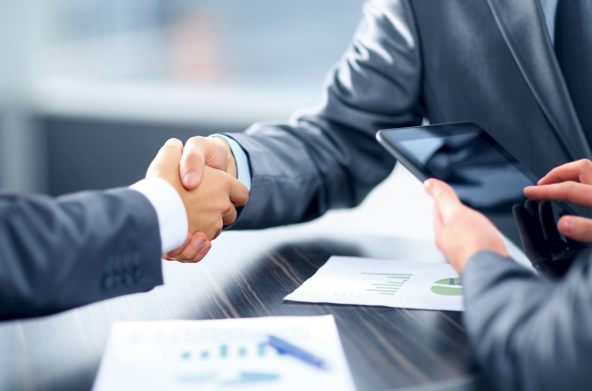 Two people in suits shaking hands over a table with documents and a tablet, suggesting a business agreement or meeting.