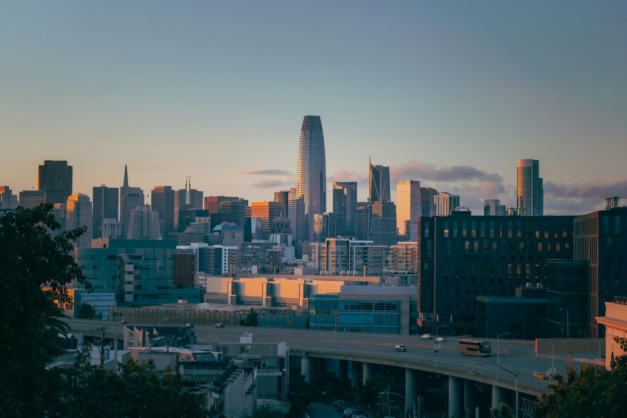 A city skyline at sunset with tall modern buildings, a prominent glass tower, and an elevated road in the foreground.