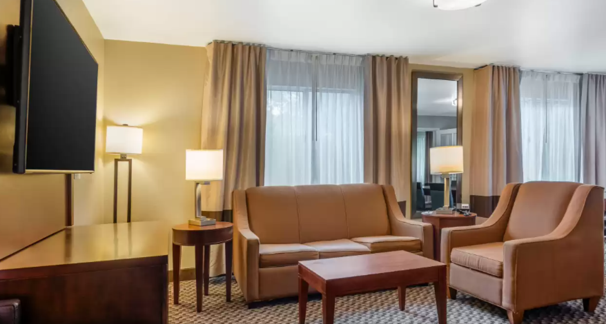 The image shows a hotel room with a sofa, armchair, coffee table, TV, and lamps, featuring large windows with curtains in a cozy setting.