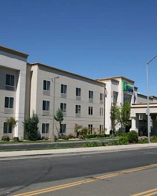 The image shows a modern, three-story hotel building with a driveway and a clear blue sky in the background.