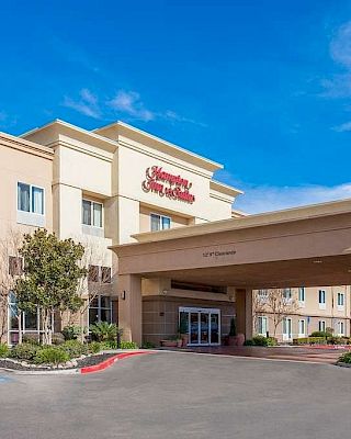 The image shows the exterior of a Hampton Inn & Suites hotel, featuring a welcoming entrance with a covered driveway and clear blue sky.