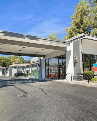 The image shows the entrance of a building with an awning, surrounded by trees and a driveway, with signage on the right side of the structure.