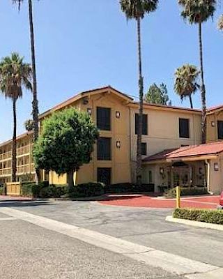 A two-story hotel building with a red-tiled roof, palm trees, and parked cars along the driveway in a sunny setting.