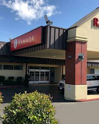 The image shows the entrance of a Ramada hotel, featuring a sign and a portico with a van parked in front.