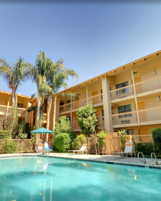 A swimming pool surrounded by palm trees and multi-story buildings with balconies, featuring umbrellas and lounge chairs in a sunny setting.