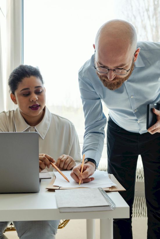 Two people are working together at a desk. One is seated with a laptop, and the other, standing, is writing on a notepad.