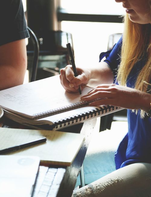 A group of people having a meeting at a table with a laptop, notebooks, and a mug. One person is taking notes with a pen.