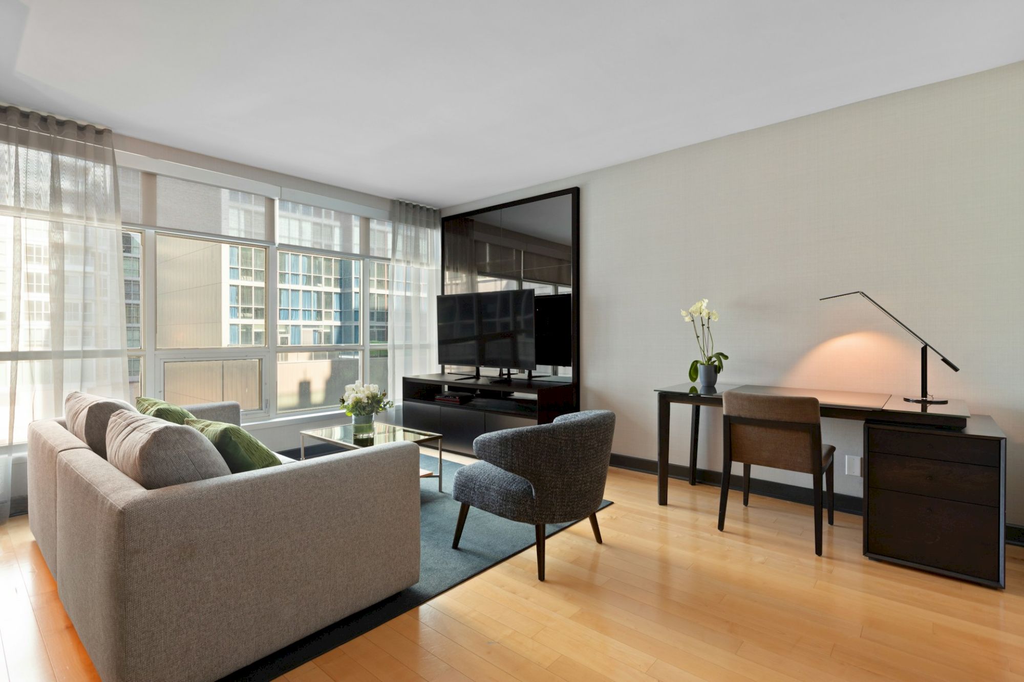 A modern living room with a sofa, armchair, TV, desk, chair, floor lamp, and large windows offering city views.